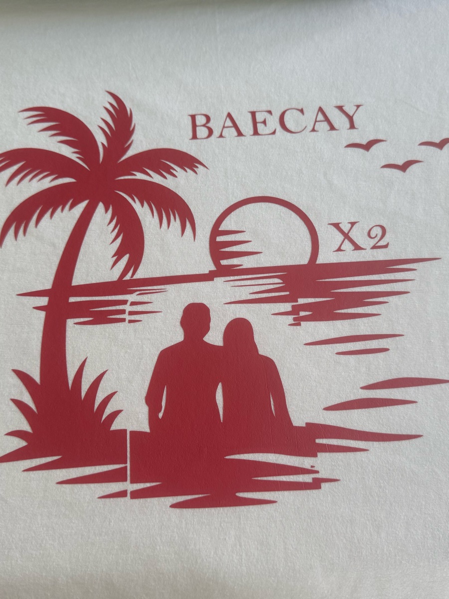 A white tee printed in deep red with BAECAY x2, a palm tree and a couple sitting together facing a setting sun.