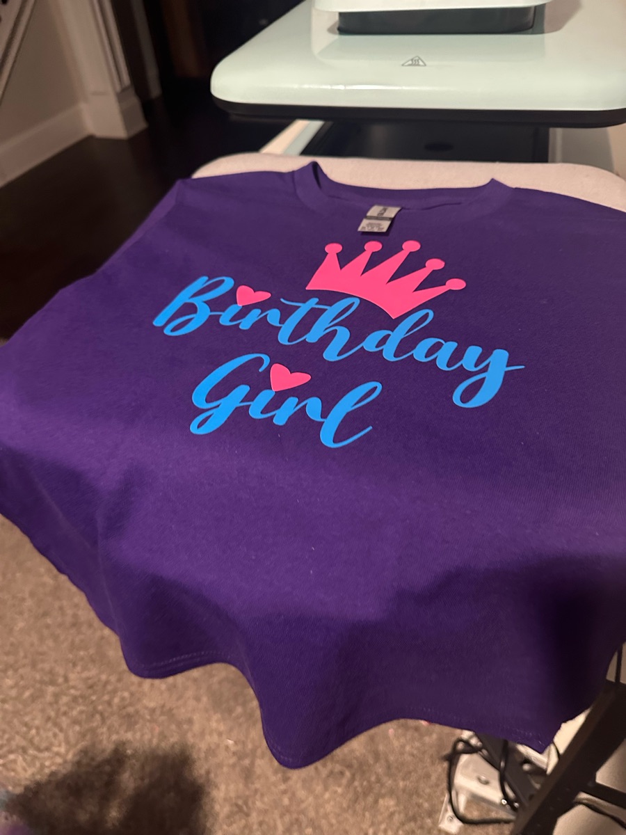 A purple tee fresh off the heat press, printed birthday girl in blue script under a pink crown.