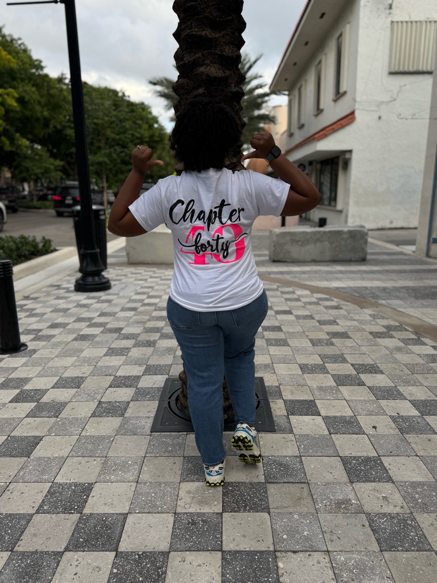 The chapter forty tee worn on the birthday trip, seen from behind on a palm-lined street with both arms raised.