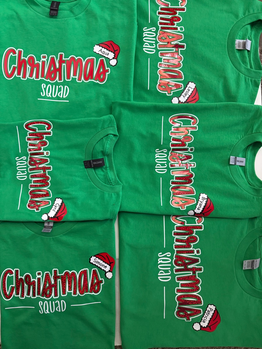 Green tees laid out in rows, each printed Christmas squad in red and white, with a Santa hat carrying one person's name.