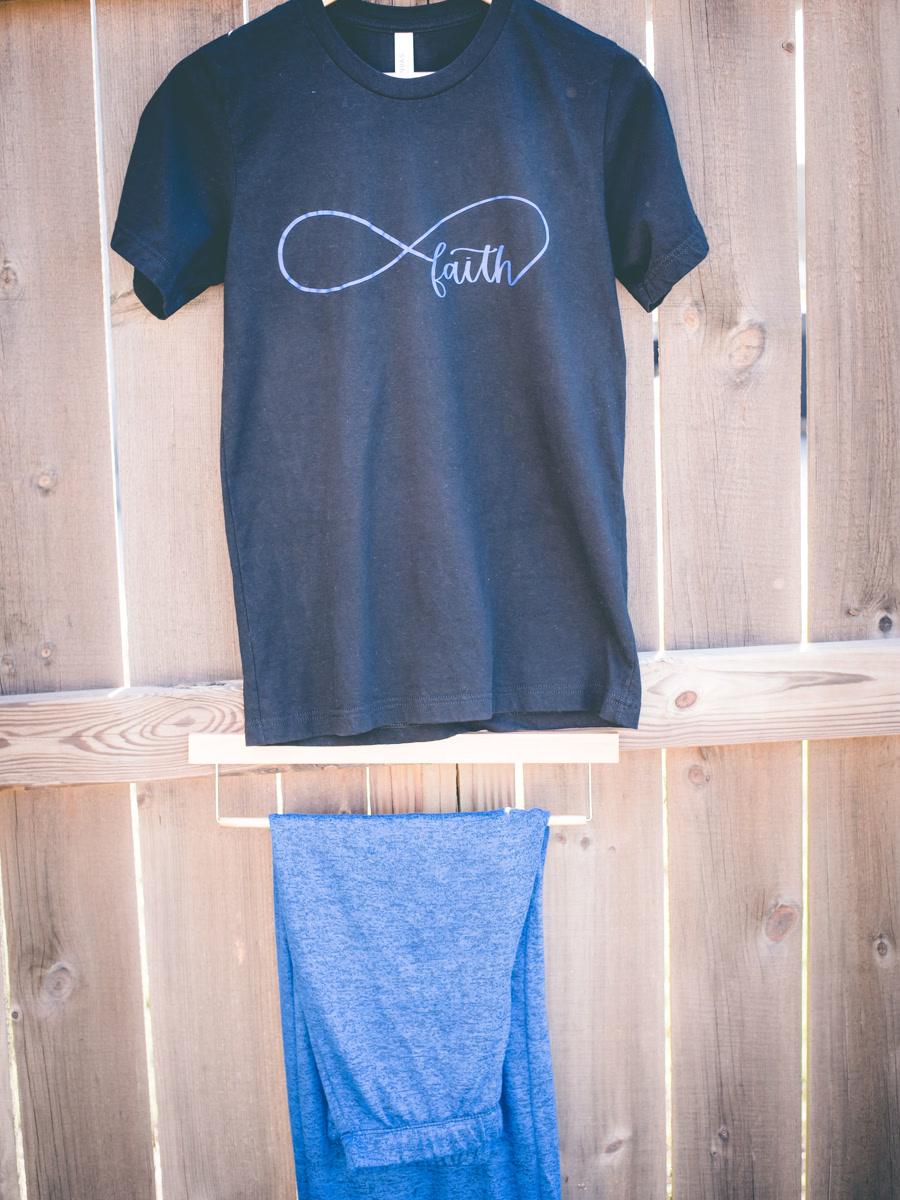 A navy tee printed with an infinity symbol drawn into the word faith, hanging from a wooden rail.