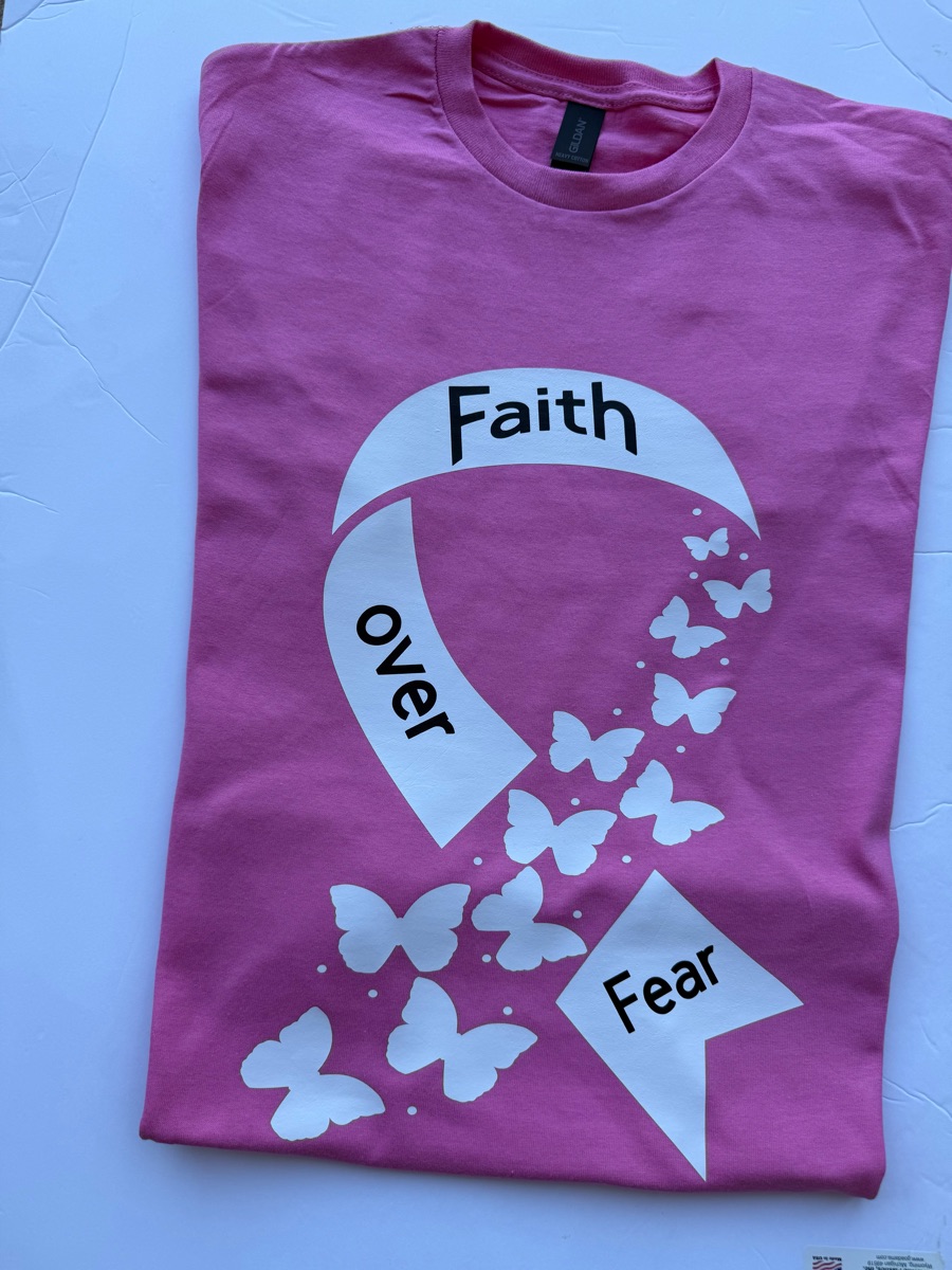 A magenta tee printed with a white awareness ribbon reading faith over fear, trailing a cloud of white butterflies.