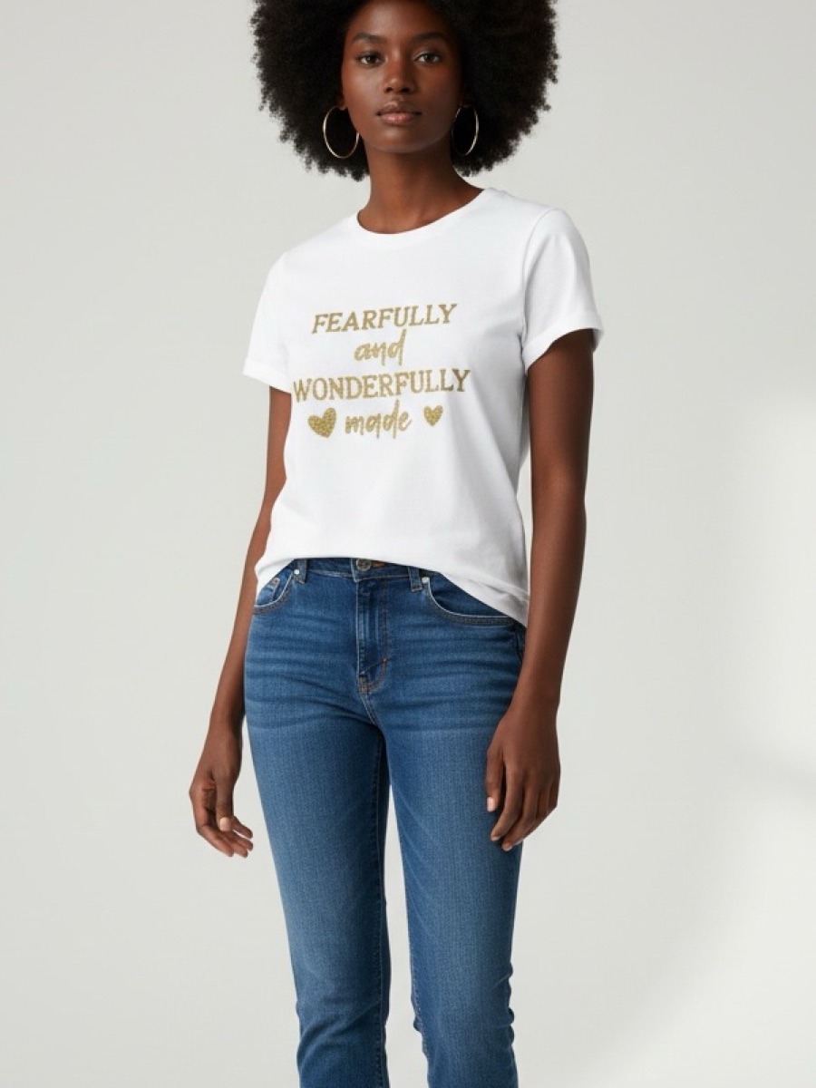 A woman in a white tee printed fearfully and wonderfully made in gold, worn with jeans and canvas sneakers.