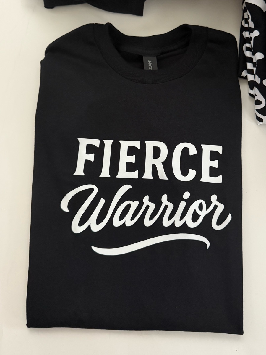 A black tee printed FIERCE in white block letters over warrior in script, folded on a table with more of the same order behind it.