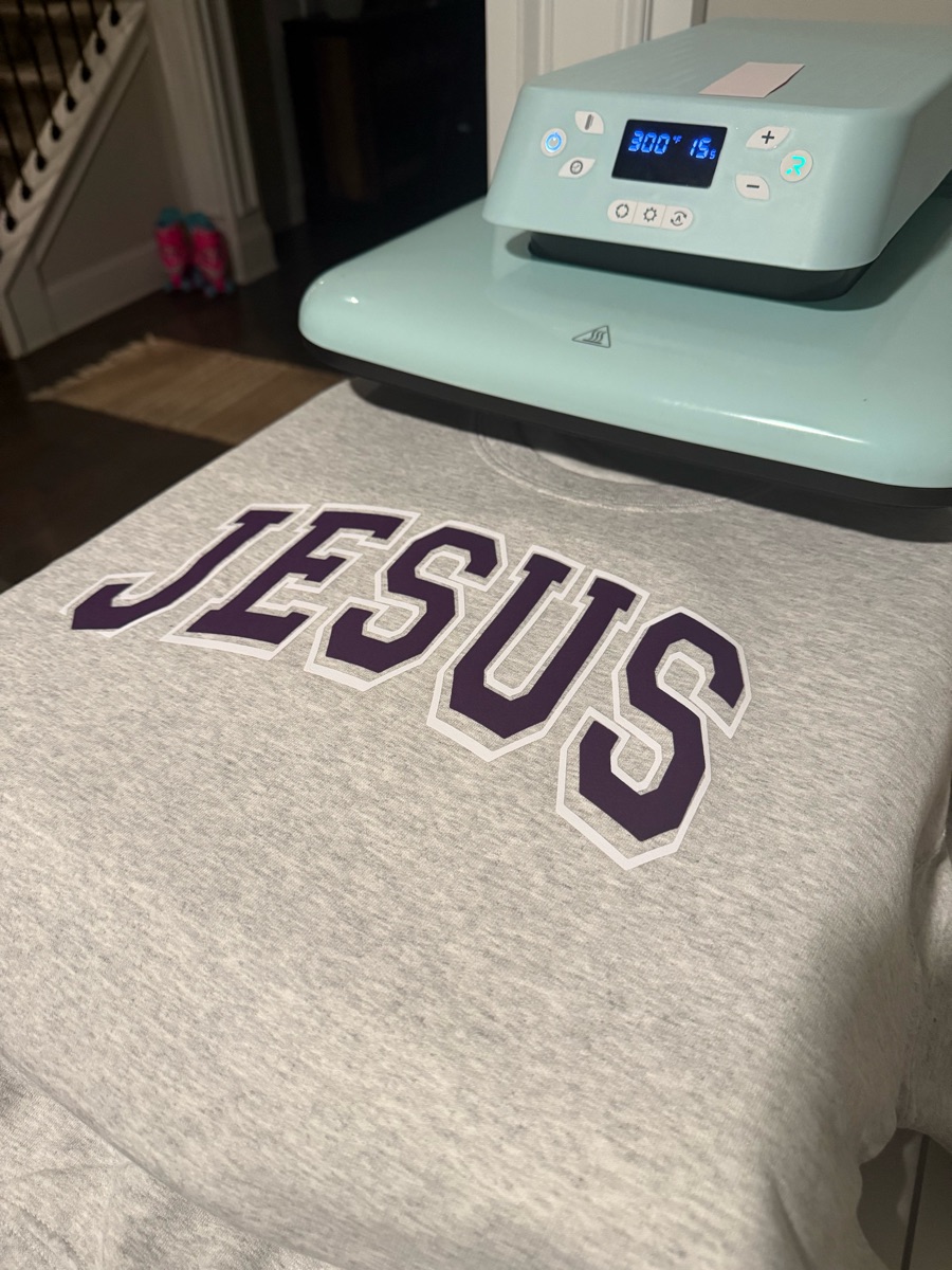 A heather grey sweatshirt on the heat press, printed JESUS in purple varsity letters outlined in white.