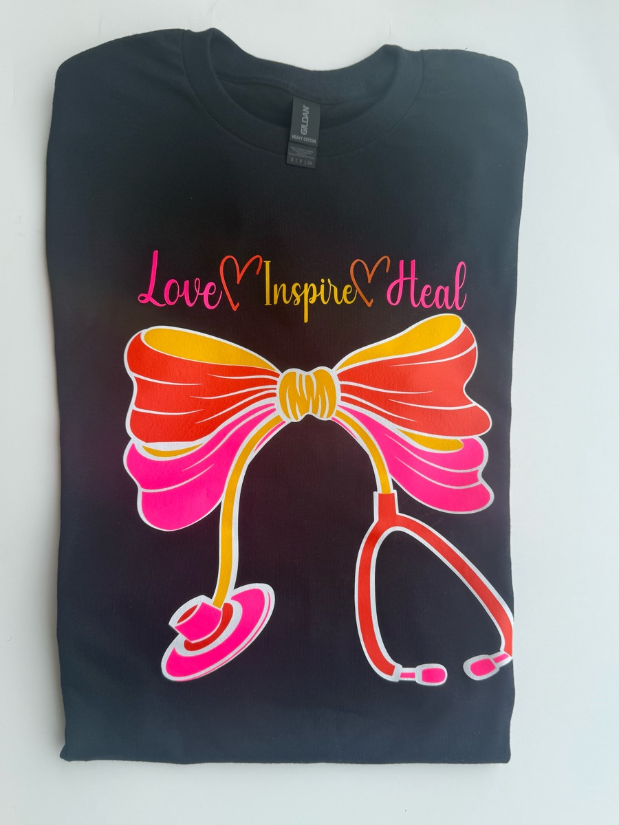 A black tee printed with love, inspire, heal in pink script above a big pink and orange bow tied from a stethoscope.