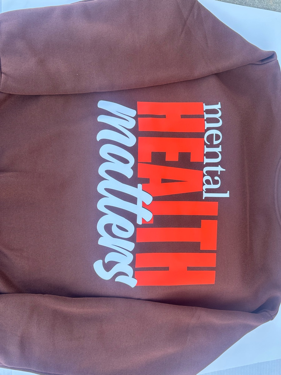 The back of a brown hoodie printed mental HEALTH matters, with HEALTH in tall orange-red letters and matters in pale blue script.