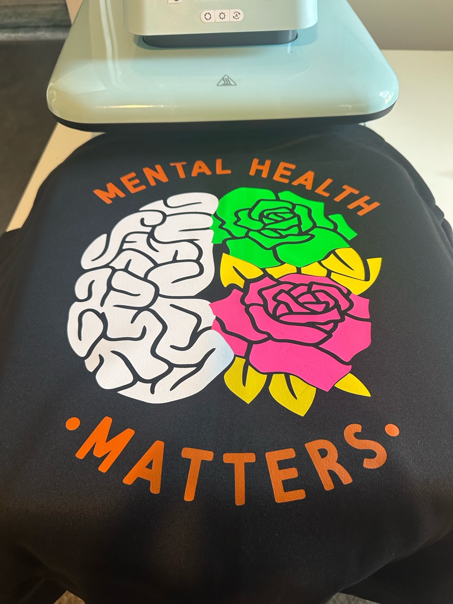 A black sweatshirt on the heat press, printed mental health matters around half a white brain and half bright green, pink and yellow roses.