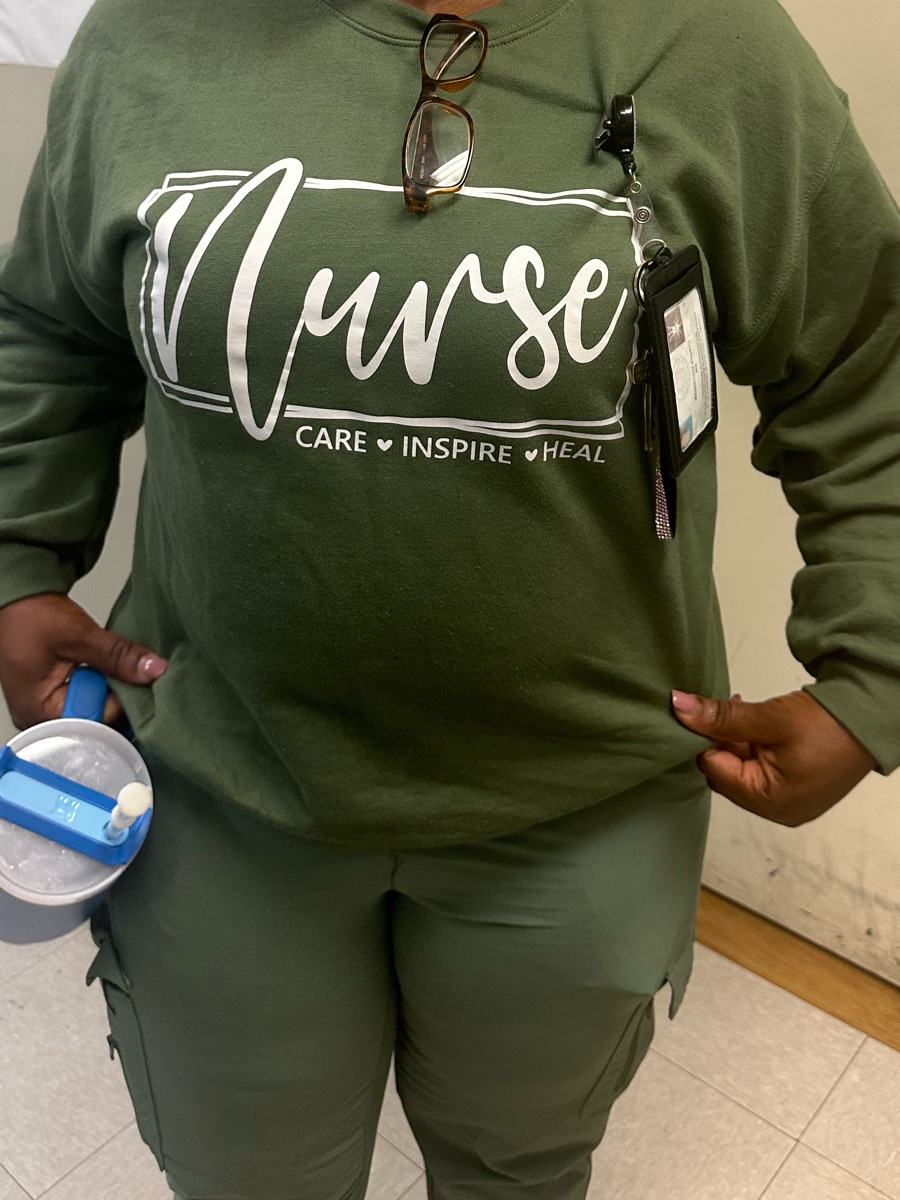 A forest green sweatshirt printed Nurse in white script with care, inspire, heal underneath, worn at work with matching green pants.