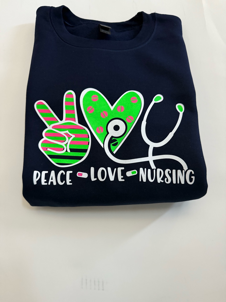 A navy sweatshirt printed peace, love, nursing under a pink and green peace sign, a green heart and a white stethoscope.
