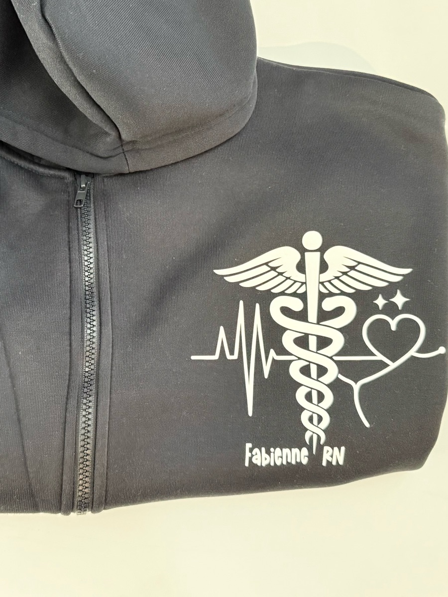 A charcoal zip hoodie with a white caduceus, heartbeat line and heart on the chest, and the wearer's first name and RN underneath.
