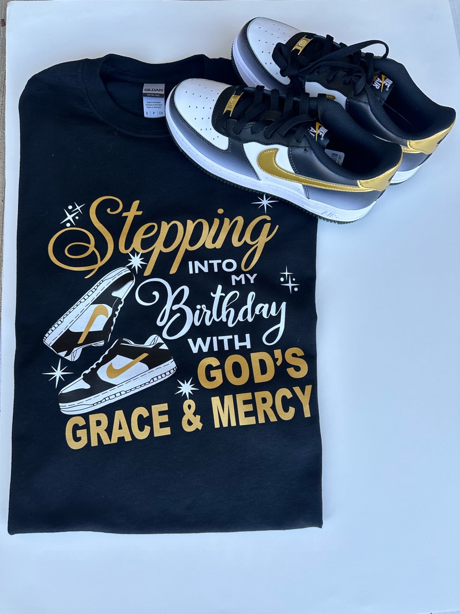 A black tee printed stepping into my birthday with God's grace and mercy in gold and white, with a pair of sneakers drawn beside it, laid next to black and gold sneakers.
