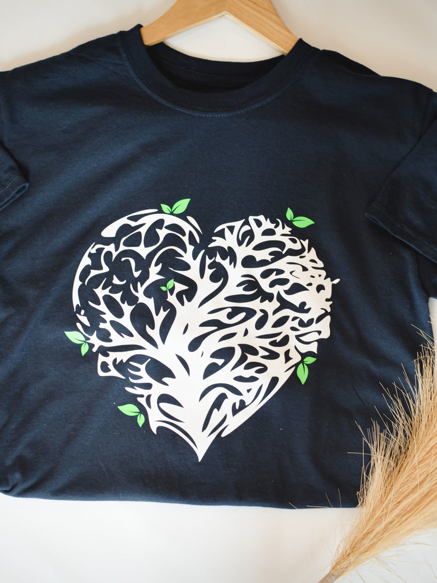 A black tee printed with a white heart made of tree branches, laid flat beside pampas grass.