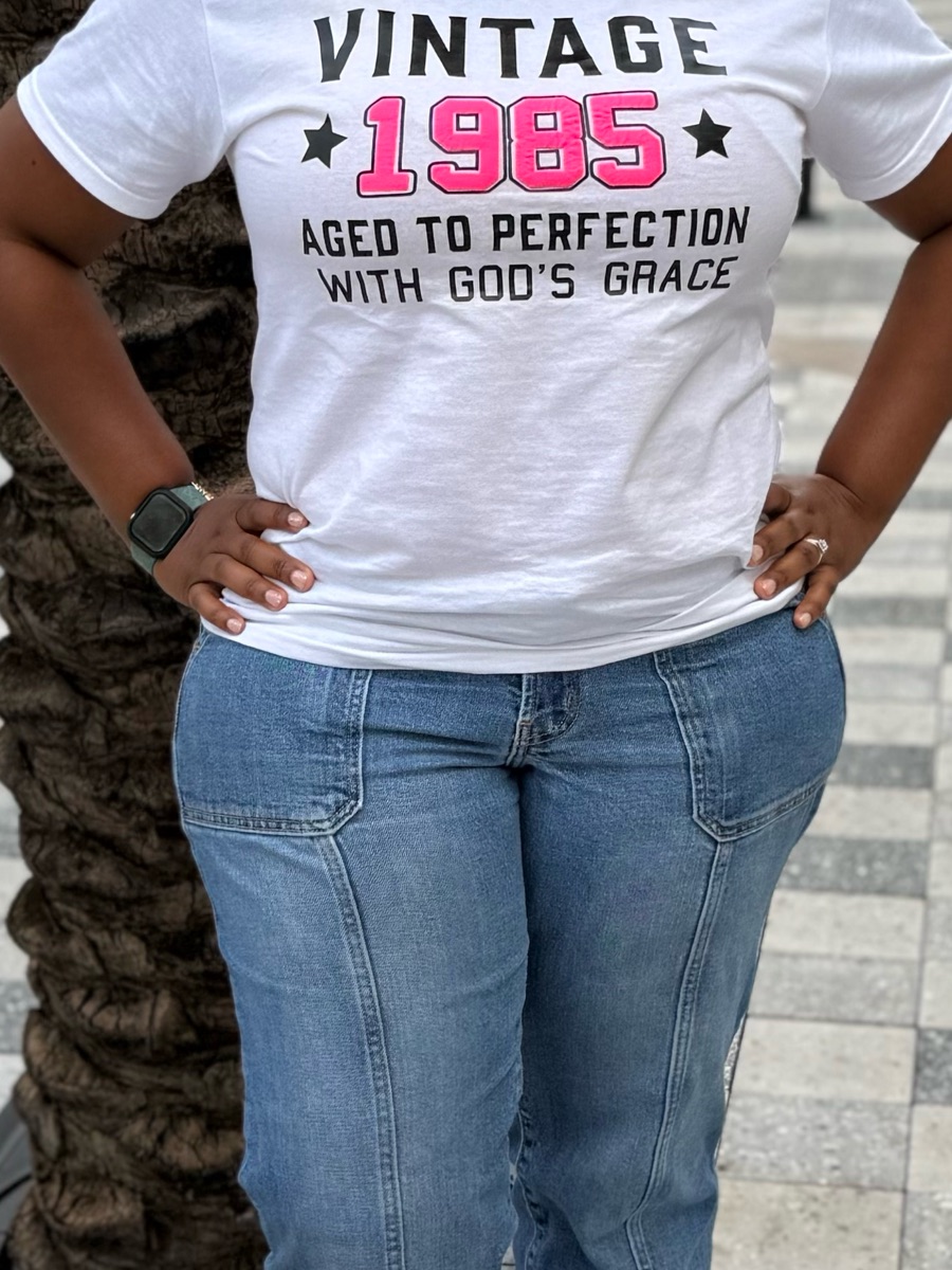 A white tee printed vintage 1985, aged to perfection with God's grace, worn with jeans and hands on hips.