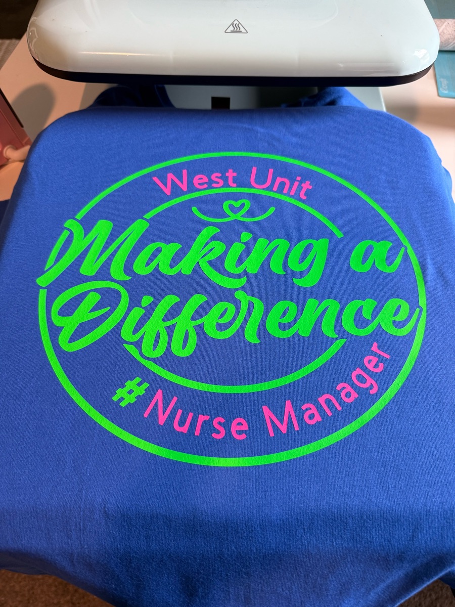 A royal blue tee on the heat press, printed West Unit, making a difference, #nurse manager in neon green and pink inside a circle.